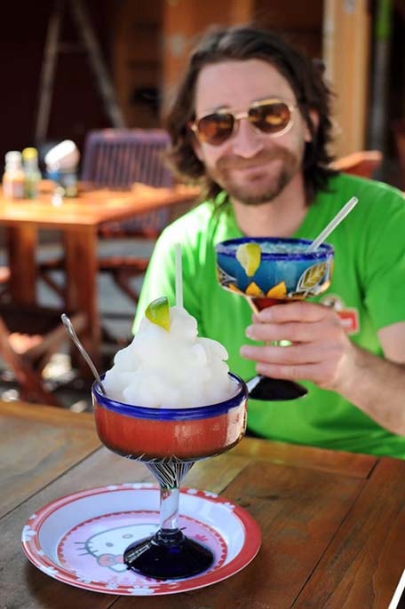 Large frozen margarita sitting on a wooden table with Randy holding up a margarita in the background
