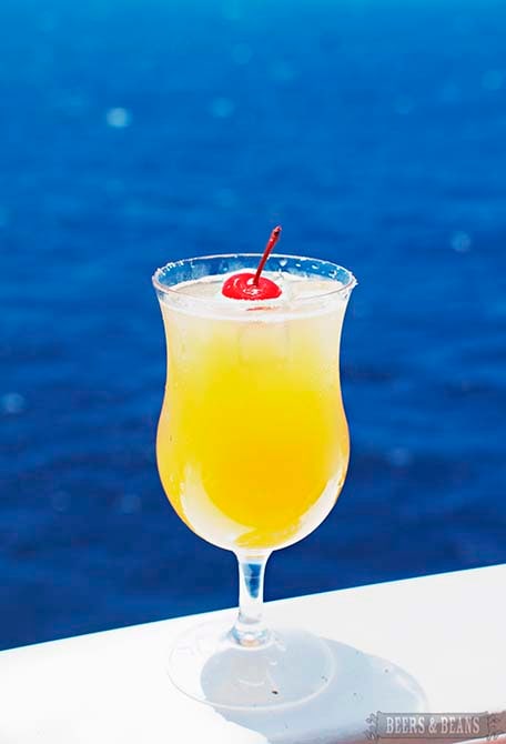 Orange drink with red cherry on top with bright blue ocean behind