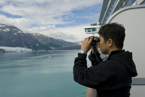 man wearing a black sweater looking at the natural beauty of alaska through binoculars on a cruise balcony