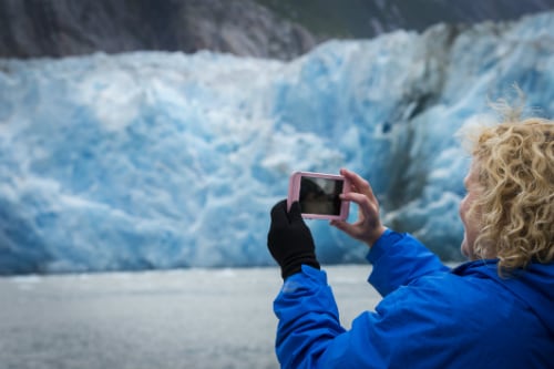 woman wearing blue jacket, taking a picture of glaciers through her pink smart phone